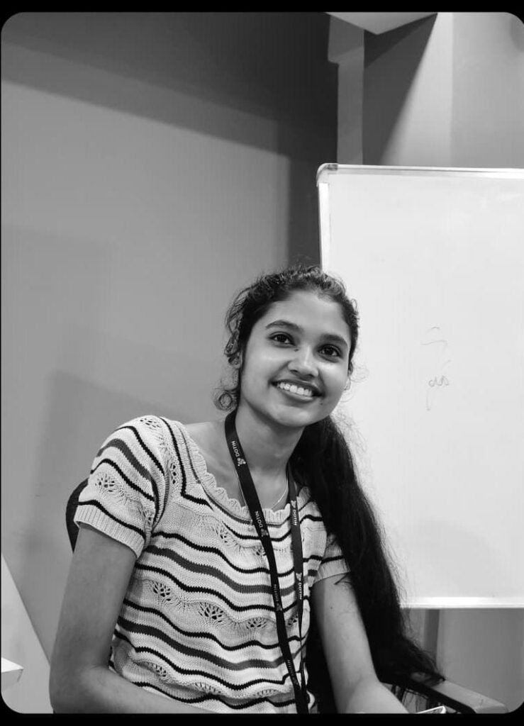 A black and white photograph captures a young woman with a bright smile, seated in what appears to be an office or classroom setting. She has long, dark hair draped over one shoulder and is wearing a striped, short-sleeved top along with a lanyard around her neck. Behind her, a plain wall and a white whiteboard with some faint writing are visible, suggesting a professional or educational environment. The lighting is soft, highlighting her expression and creating a clean, high-contrast aesthetic typical of monochrome photography.